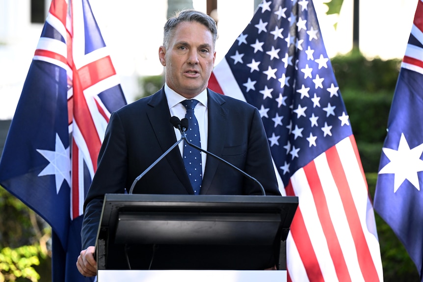 A middle-aged man in dark suit speaks at podium in front of Australian and US flags