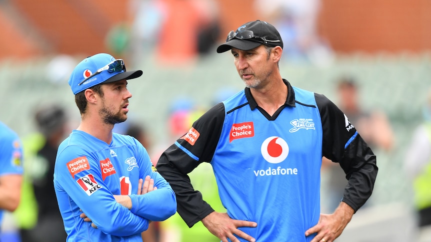 Two men in blue cricket uniforms stand side-by-side, talking.