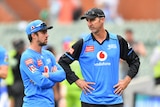 Two men in blue cricket uniforms stand side-by-side, talking.