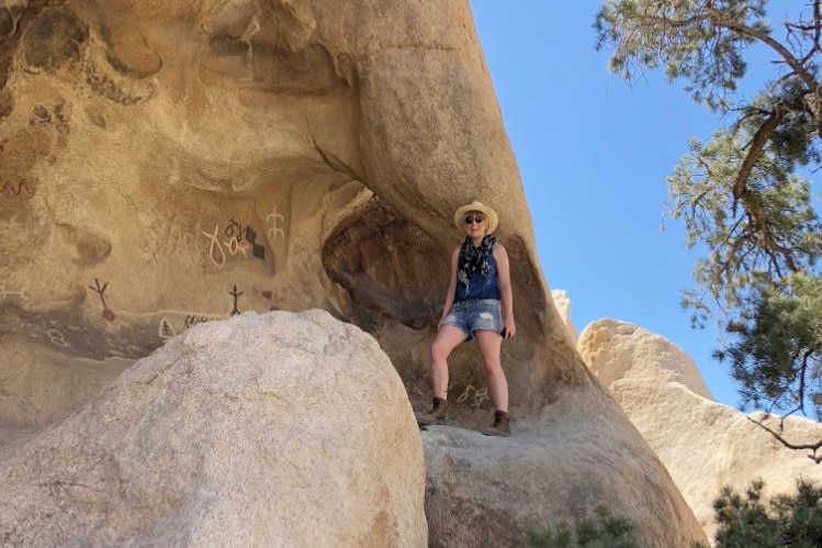 A woman in sunglasses, hat, singlet and boots stands smiling, surrounded by large boulders, with blue sky above.