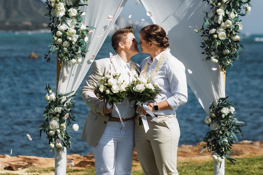 Sarah and Jess Jonassen stand in front of a wedding arbour cover in flowers. They hold bouquets and kiss.