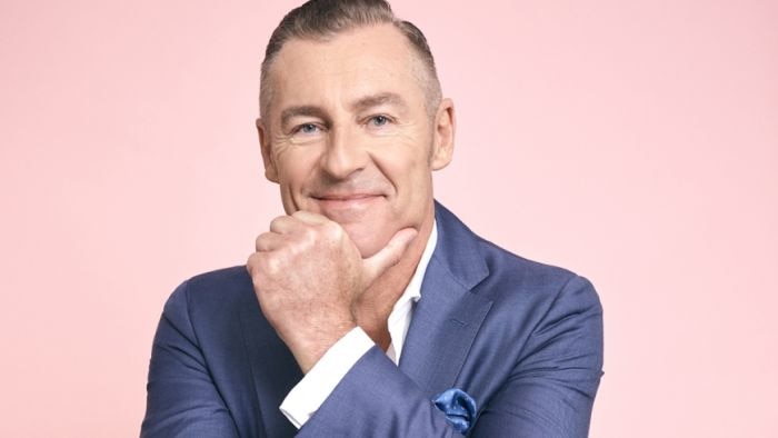 Paul Dyer smiles with his hand to his chin, against a pink background