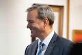 Smiling man in a suit, stands holding the budget paper for 2023 