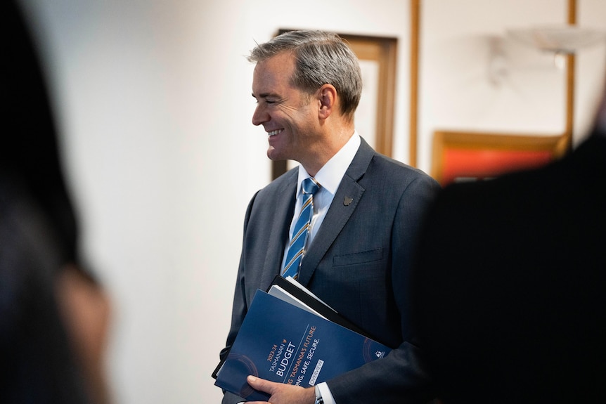 Smiling man in a suit, stands holding the budget paper for 2023 