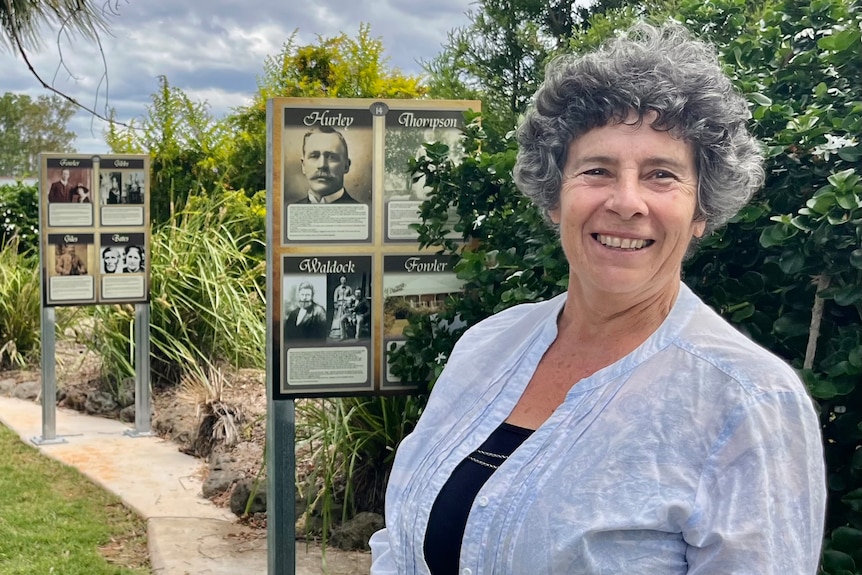 A woman with grey hair stands in front of a sign which outlines the history of pioneer families. 