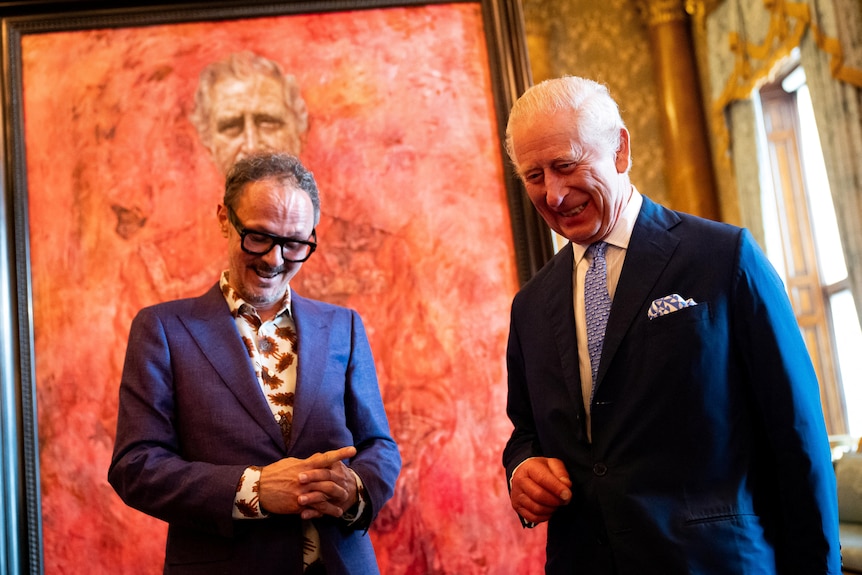 Two men wearing suits looking down and laughing with a big red painting behind them