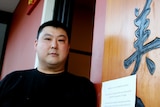 A man with dark hair stands outside a restaurant with Chinese writing on sign
