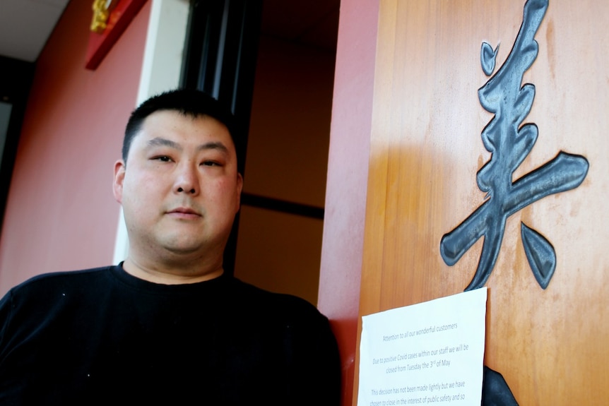 A man with dark hair stands outside a restaurant with Chinese writing on sign