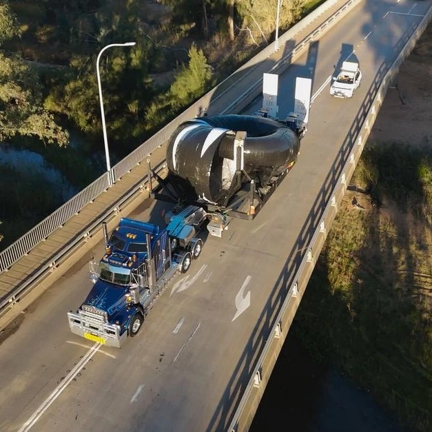 A flat bed truck, carrying a large metal sculpture, and follwed by an escort car, crosses a bridge 