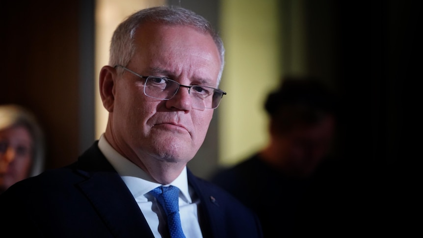 Scott Morrison wearing a blue tie and glasses looks to the left of camera in a dark room