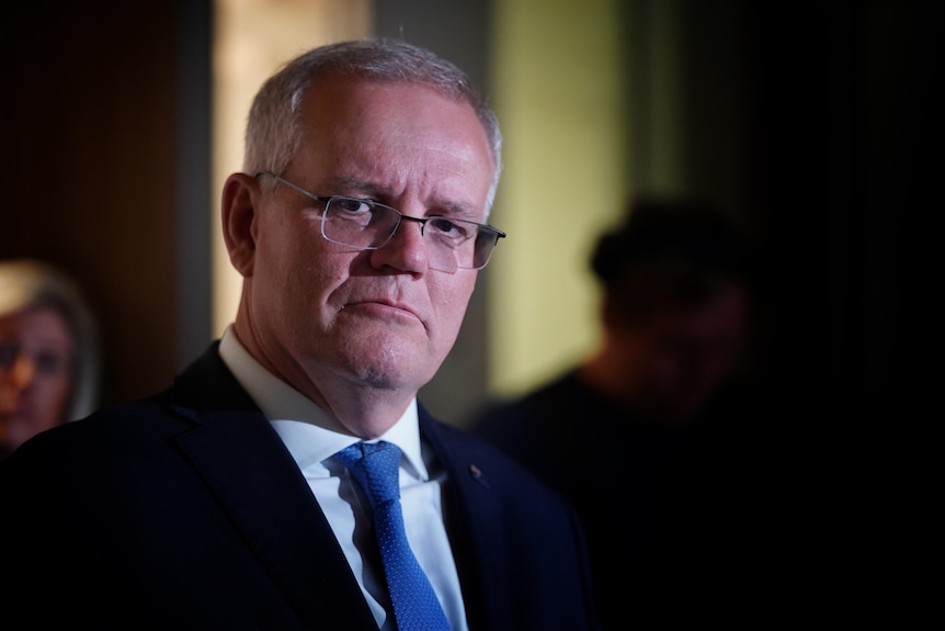 Scott Morrison wearing a blue tie and glasses looks to the left of camera in a dark room