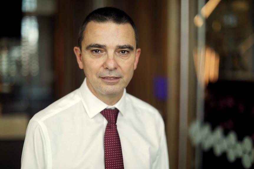 A man wearing a shirt and tie, stands indoors looking at the camera with a neutral expression.