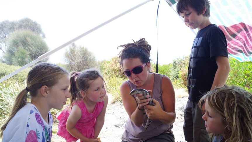 A woman holds a lizard as four children look on.