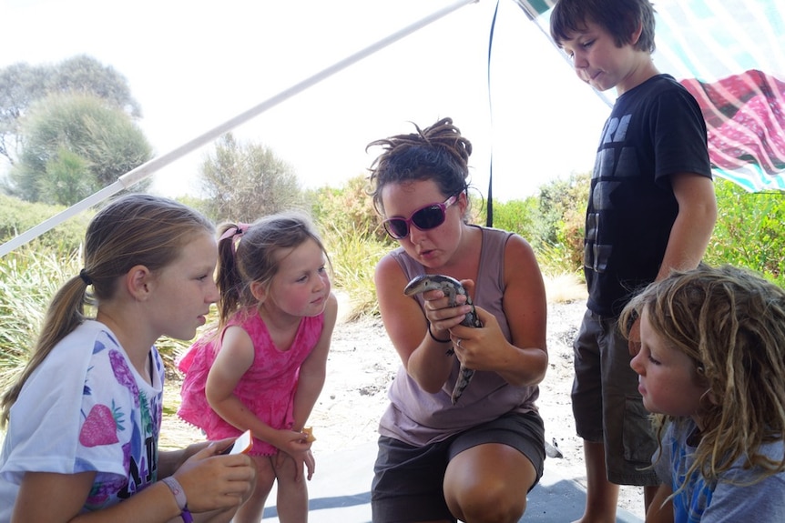 A woman holds a lizard as four children look on.