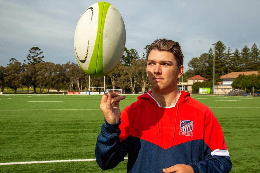 Man in his early twenties stands on a field and throws a rugby ball up into the air.