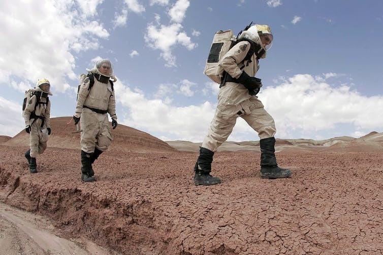 Three people in spacesuits walk through a desert