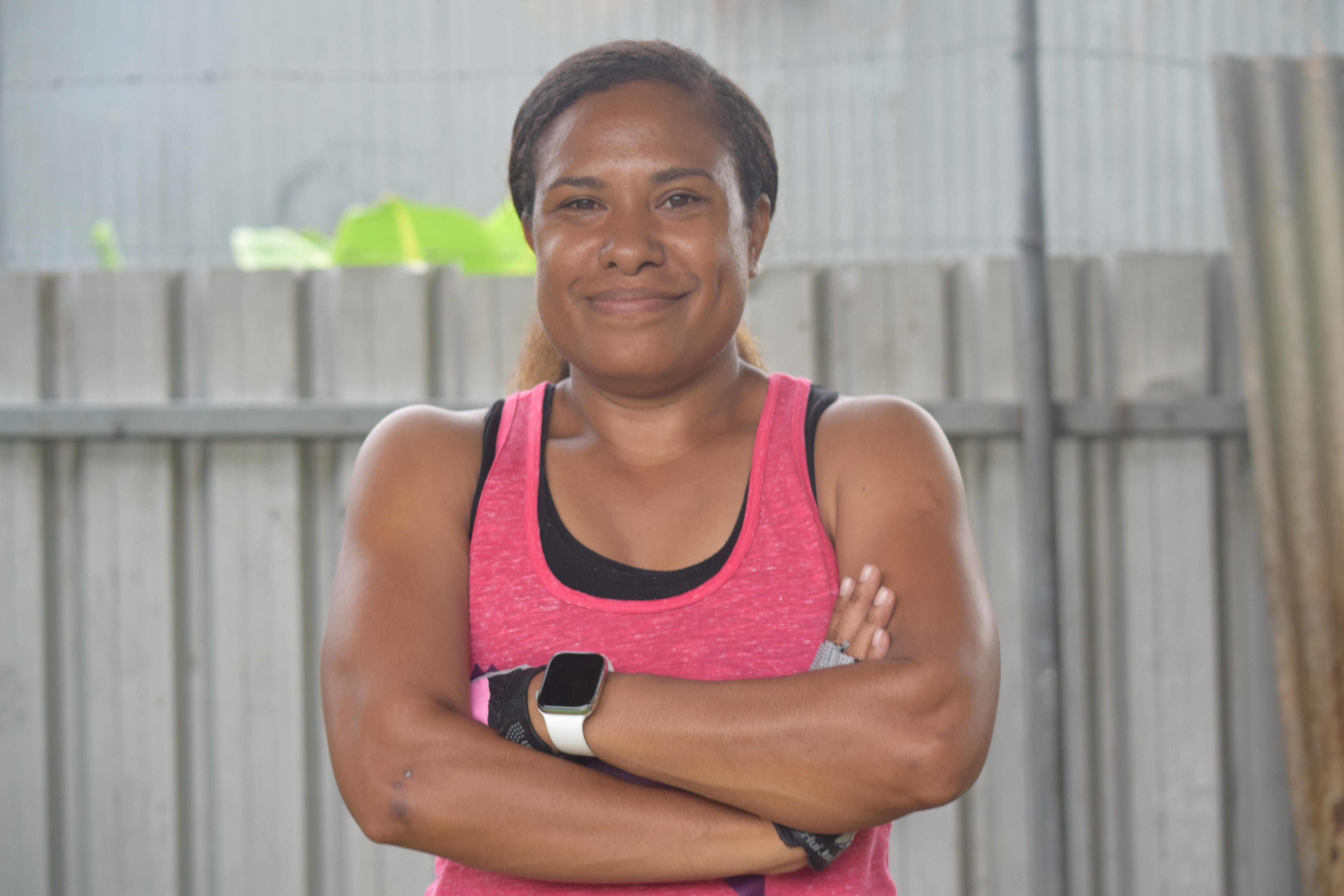 A Papua New Guinean woman wearing a pink singlet has her arms crossed and smiles at the camera.
