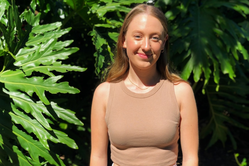 A young woman stands and looks to the camera while smiling, in front of green philodendron plants. She wears a brown singlet.