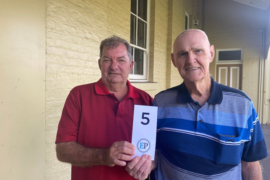 man in red shirt stands next to elderly man in blue stripy shirt holding a card with the number 5 written on it