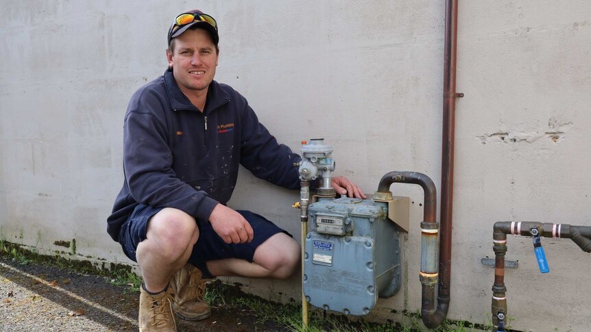 A man in a worn navy jumper and a hat and sunnies on his head crouches down next to a gas meter.