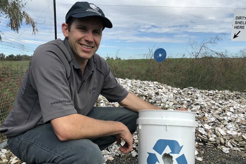 A man wearing a grey shirt and a black cap crouches in front of a big pile of used oyster shells.