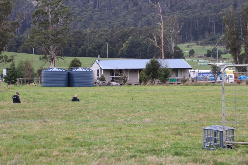 Two police officers walk through a paddock towards a rural property.