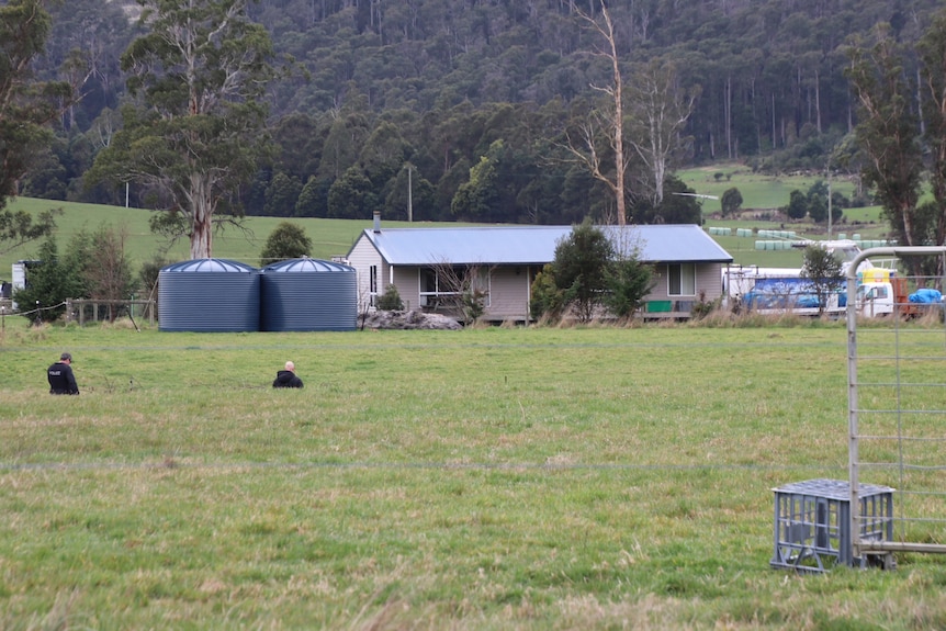 Two police officers walk through a paddock towards a rural property.