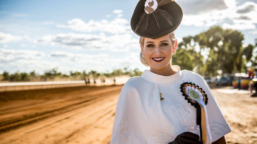 A beautifully dressed young lady stands in front of a red dirt track with horses racing behind.