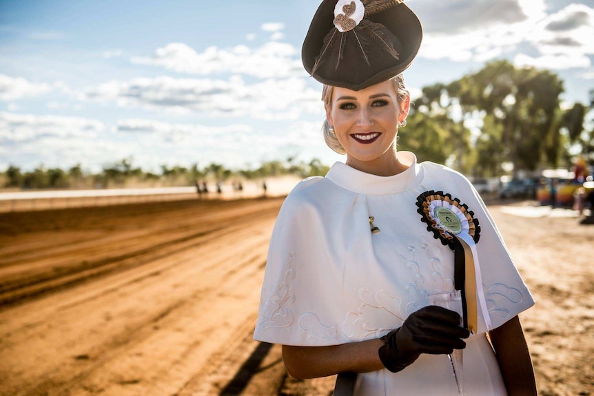 A beautifully dressed young woman stands in front of a red dirt track with horses racing behind.