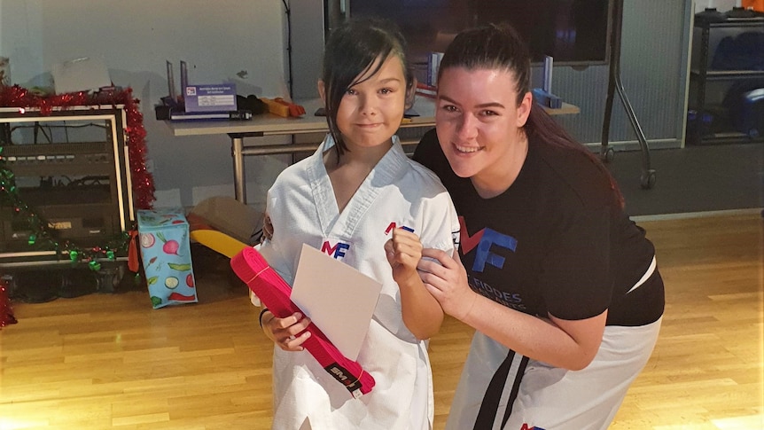 Child wears martial arts uniform while holding red belt and certificate, standing next to her mum
