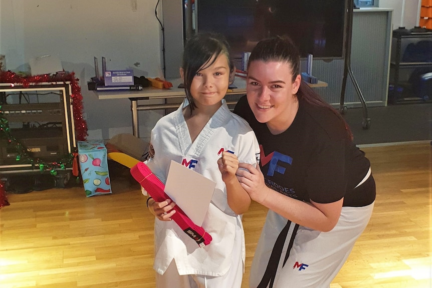 Child wears martial arts uniform while holding red belt and certificate, standing next to her mum