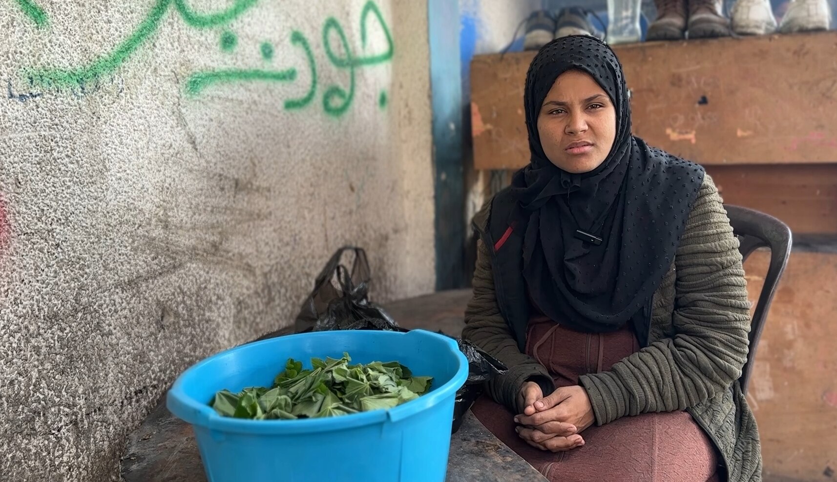A woman wearing a dark blue hijab sits in a chair leaning over a table with a bowl full of greens.