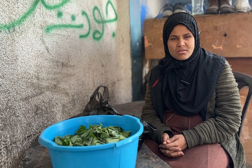 A woman wearing a dark blue hijab sits in a chair leaning over a table with a bowl full of greens.