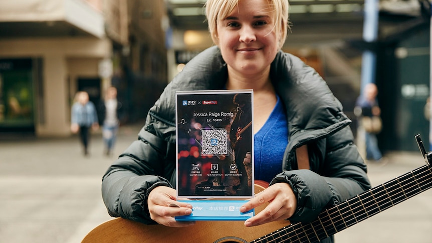 A woman with a guitar holds a sign with a QR code on it. A QR code is like a barcode you scan with your phone.