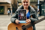 A woman with a guitar holds a sign with a QR code on it. A QR code is like a barcode you scan with your phone.