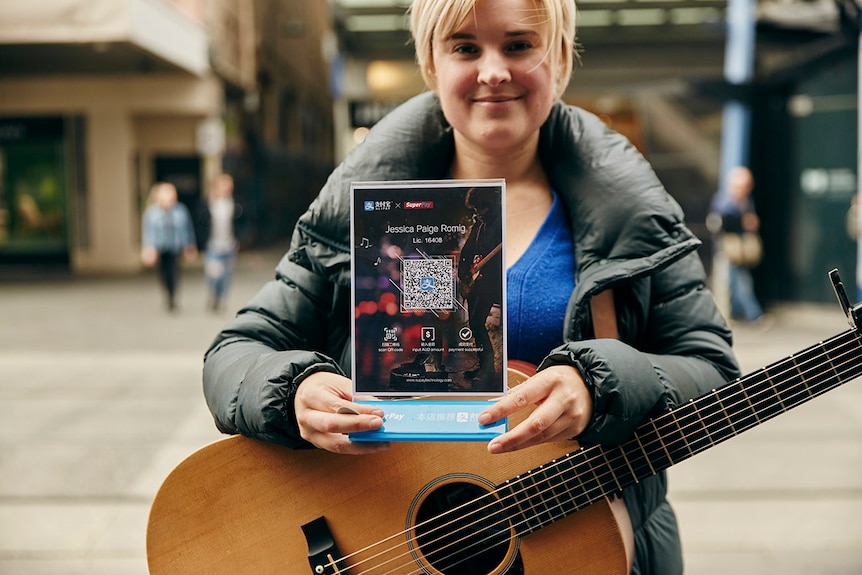 A woman with a guitar holds a sign with a QR code on it. A QR code is like a barcode you scan with your phone.