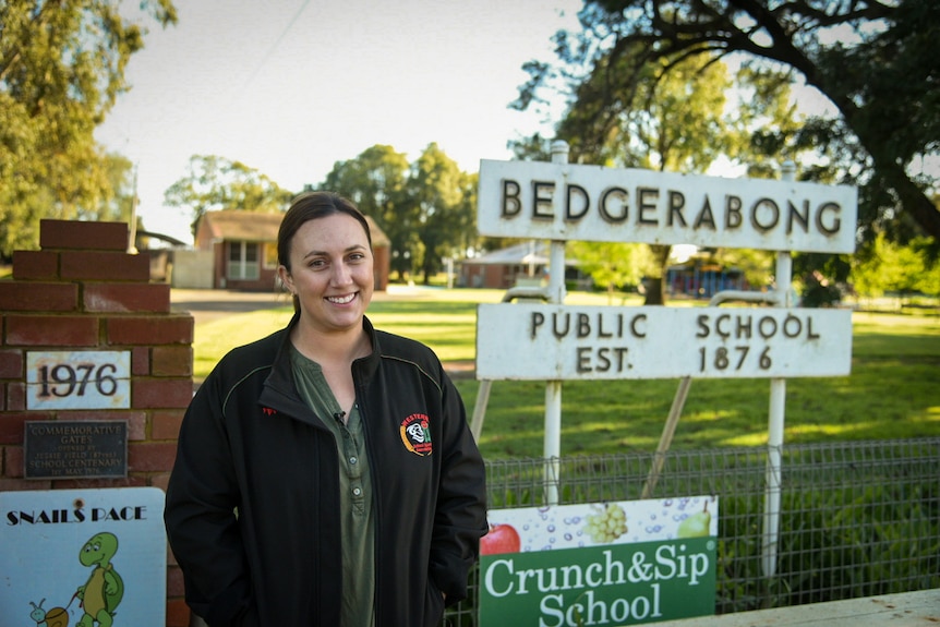 A woman wearing a black jacket smiles in front of a sign that says "Bedgerabong Public School".