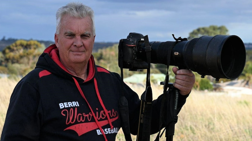 A white man with grey hair wearing a navy and red jumper and holding a big camera in a bushy landscape. He looks proud.