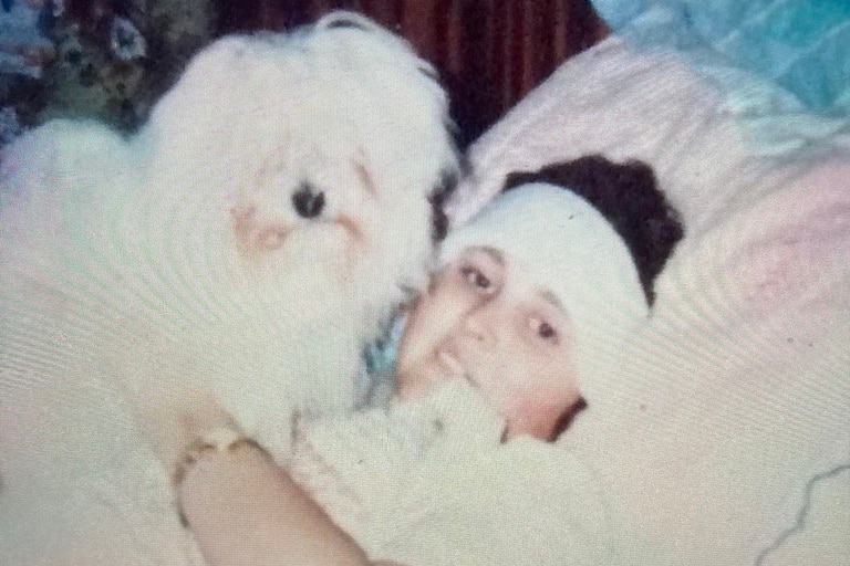 Young girl with a fluffy dog, laying in a white bed with a bandage around her head.