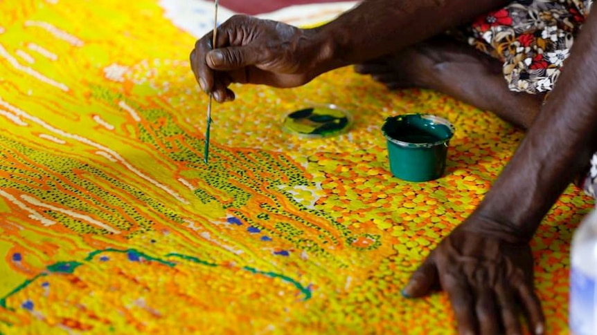 The hands of an indigenous woman hold a stick with green paint on as she applies it to a colourful aboriginal painting