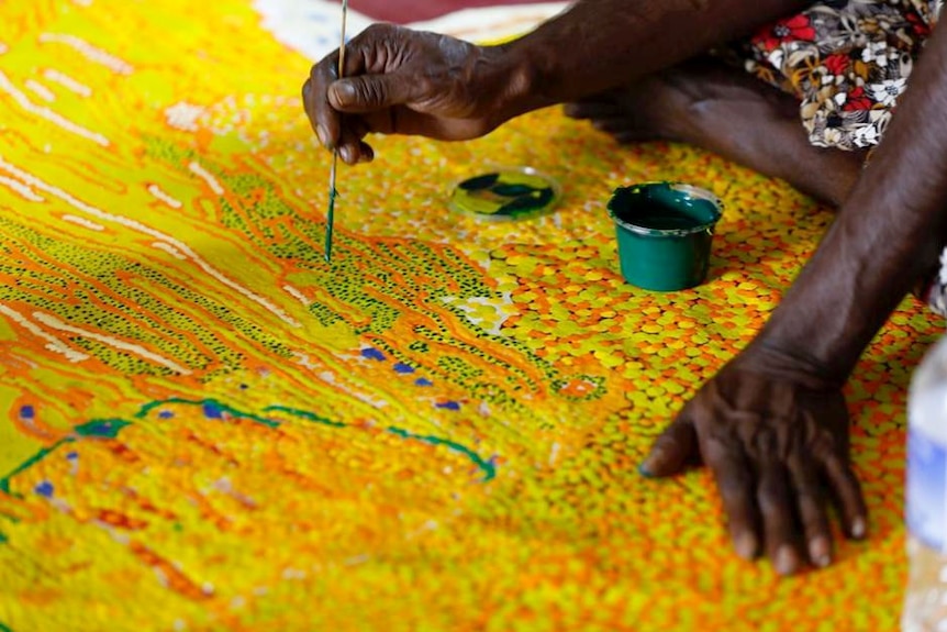 The hands of an indigenous woman applying paint to a colourful aboriginal painting, the Kalyu.