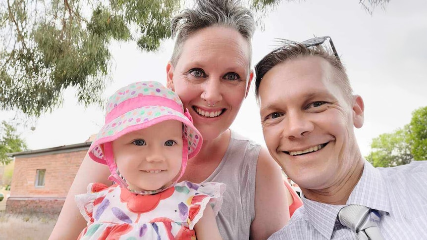 A woman with short hair holds her toddler daughter and poses for a selfie with her partner and father of their child.