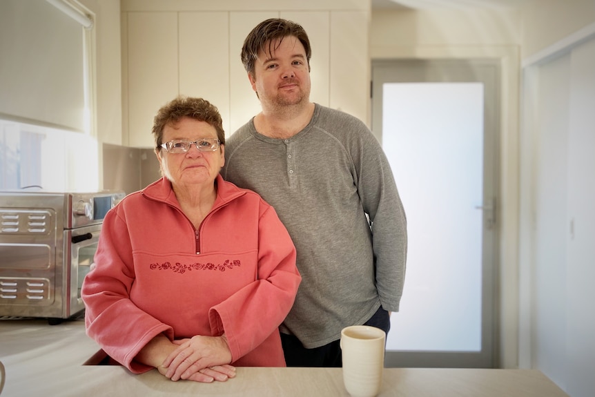 A mother and her middle aged son stand proudly in their clean, white kitchen.