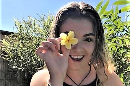 A young girl smiling at the camera holding a frangipani flower.