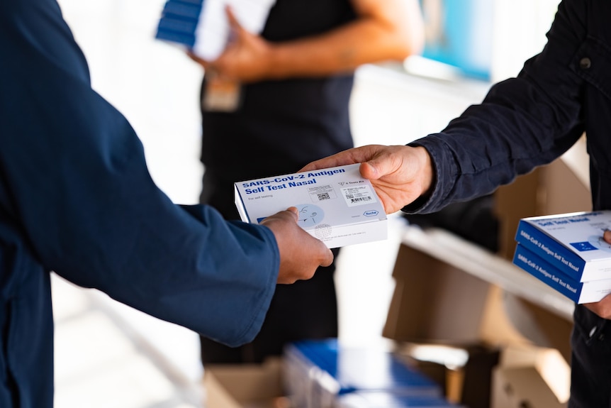 A small box with a rapid antigen test inside is passed from one man to another.