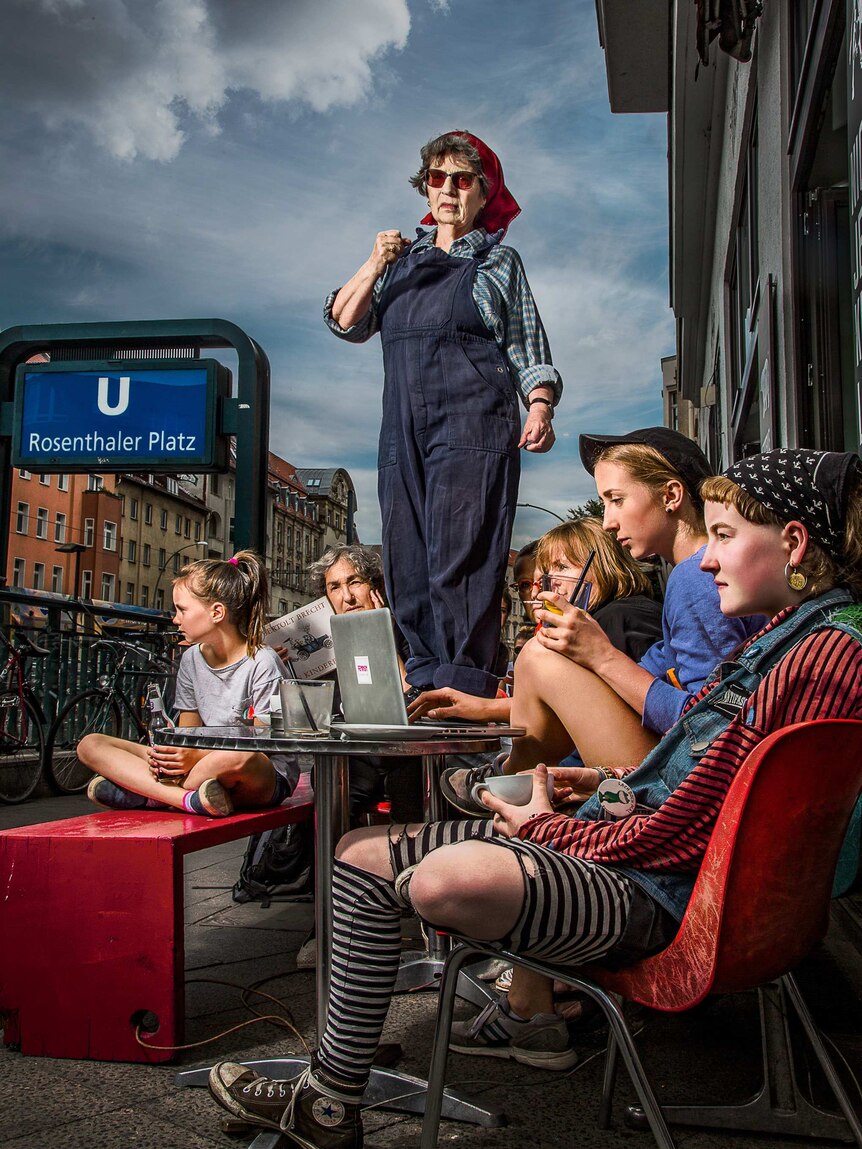 Salomea Genin, wearing overalls and a red headscarf, stands on a table in a Berlin street. A number of young women sit below her