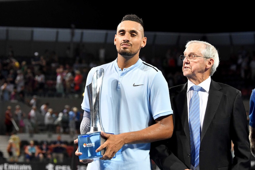 Nick Kyrgios holds the winners' trophy at the Brisbane International alongside Roy Emerson.