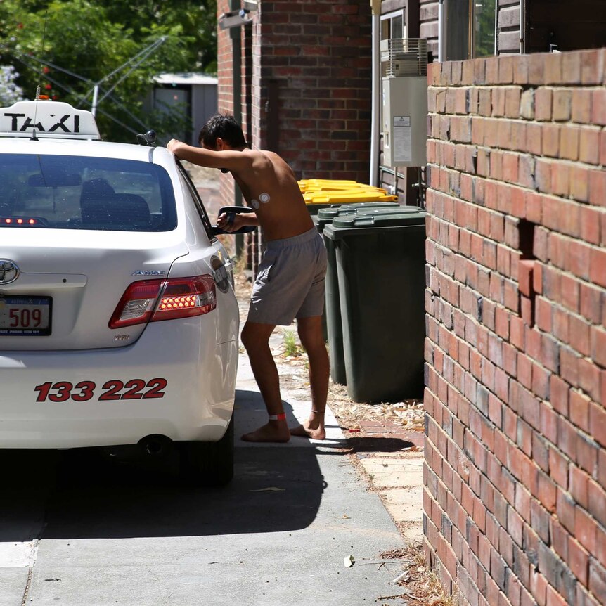 A man wearing a hospital leg band and no shirt leans on a taxi in the driveway of the house.
