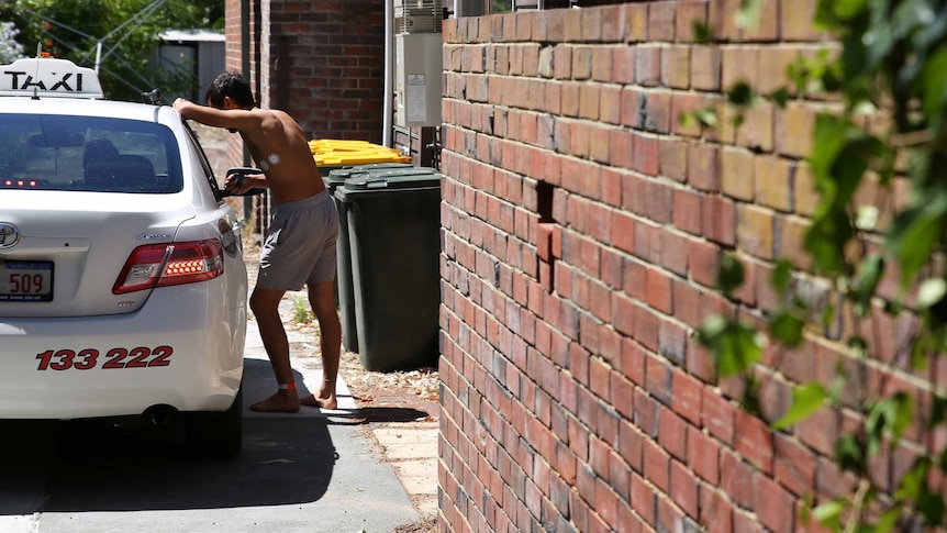 A man wearing a hospital leg band and no shirt leans on a taxi in the driveway of the house.