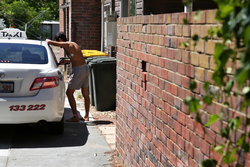 A man wearing a hospital leg band and no shirt leans on a taxi in the driveway of the house.